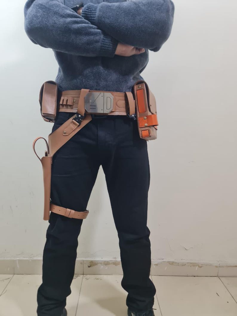 May include: A tan leather utility belt with multiple pouches and holsters, featuring a silver buckle. The belt is worn with black trousers and a grey jumper. A holster is attached to the leg.