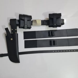 K&ouml;nnte beinhalten: Ein schwarzes Leder-Utility-G&uuml;rtel-Set mit Messerscheide, zwei Taschen und zwei zus&auml;tzlichen Riemen. Der G&uuml;rtel hat eine silberne Schnalle und wird auf wei&szlig;em Hintergrund gezeigt. Die Messerscheide befindet sich auf der linken Seite.