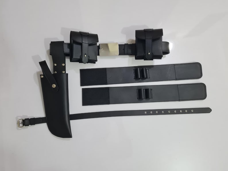 K&ouml;nnte beinhalten: Ein schwarzes Leder-Utility-G&uuml;rtel-Set mit Messerscheide, zwei Taschen und zwei zus&auml;tzlichen Riemen. Der G&uuml;rtel hat eine silberne Schnalle und wird auf wei&szlig;em Hintergrund gezeigt. Die Messerscheide befindet sich auf der linken Seite.