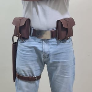 K&ouml;nnte beinhalten: Ein brauner Lederg&uuml;rtel mit gro&szlig;er silberner Schnalle, zwei Taschen und einem Holster. Das Outfit umfasst einen Oberschenkelriemen und Beinmanschetten. Die Person tr&auml;gt ein wei&szlig;es T-Shirt mit dem Aufdruck "IMPOSSIBLE".