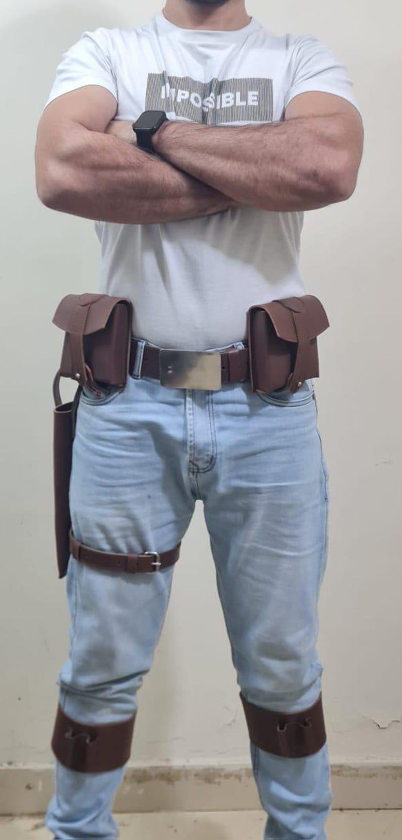K&ouml;nnte beinhalten: Ein brauner Lederg&uuml;rtel mit gro&szlig;er silberner Schnalle, zwei Taschen und einem Holster. Das Outfit umfasst einen Oberschenkelriemen und Beinmanschetten. Die Person tr&auml;gt ein wei&szlig;es T-Shirt mit dem Aufdruck "IMPOSSIBLE".
