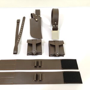 K&ouml;nnte beinhalten: Eine Sammlung brauner Lederaccessoires, darunter ein G&uuml;rtel, zwei Beutel, eine Messerscheide und zwei breite G&uuml;rtelst&uuml;cke mit Klettverschl&uuml;ssen. Die Artikel sind auf einer wei&szlig;en Oberfl&auml;che angeordnet.