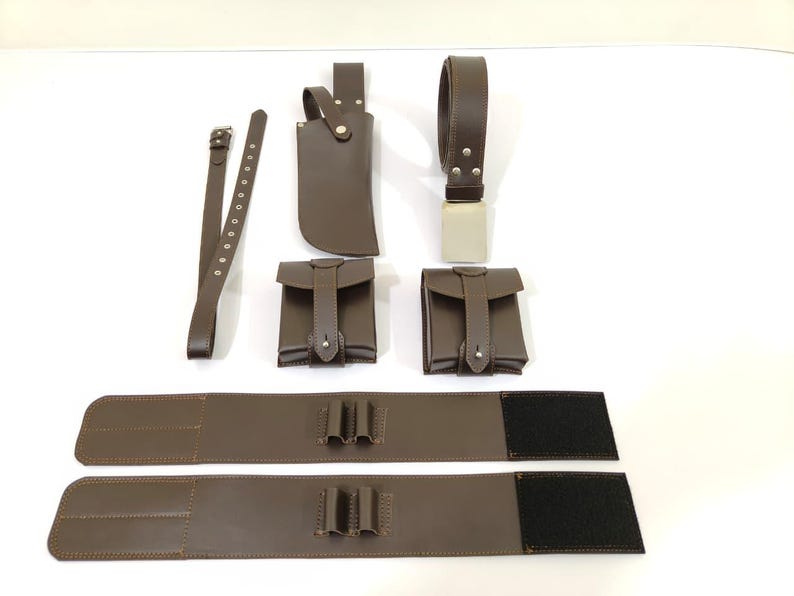 K&ouml;nnte beinhalten: Eine Sammlung brauner Lederaccessoires, darunter ein G&uuml;rtel, zwei Beutel, eine Messerscheide und zwei breite G&uuml;rtelst&uuml;cke mit Klettverschl&uuml;ssen. Die Artikel sind auf einer wei&szlig;en Oberfl&auml;che angeordnet.