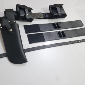 K&ouml;nnte beinhalten: Ein schwarzes Leder-Utility-G&uuml;rtel-Set mit mehreren Taschen, einer Messerscheide und zus&auml;tzlichen Riemen. Der G&uuml;rtel hat eine silberne Schnalle und ist f&uuml;r das Tragen verschiedener Gegenst&auml;nde konzipiert. Das Set liegt auf einer wei&szlig;en Oberfl&auml;che.