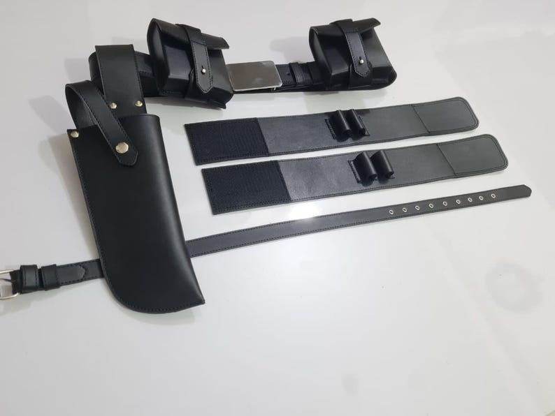 K&ouml;nnte beinhalten: Ein schwarzes Leder-Utility-G&uuml;rtel-Set mit mehreren Taschen, einer Messerscheide und zus&auml;tzlichen Riemen. Der G&uuml;rtel hat eine silberne Schnalle und ist f&uuml;r das Tragen verschiedener Gegenst&auml;nde konzipiert. Das Set liegt auf einer wei&szlig;en Oberfl&auml;che.