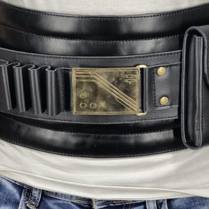 May include: Black leather belt with a gold-colored rectangular buckle. The belt features multiple loops and a small pouch. The buckle has a geometric design with three diagonal lines and three circles. The belt is worn over a white shirt and blue jeans.