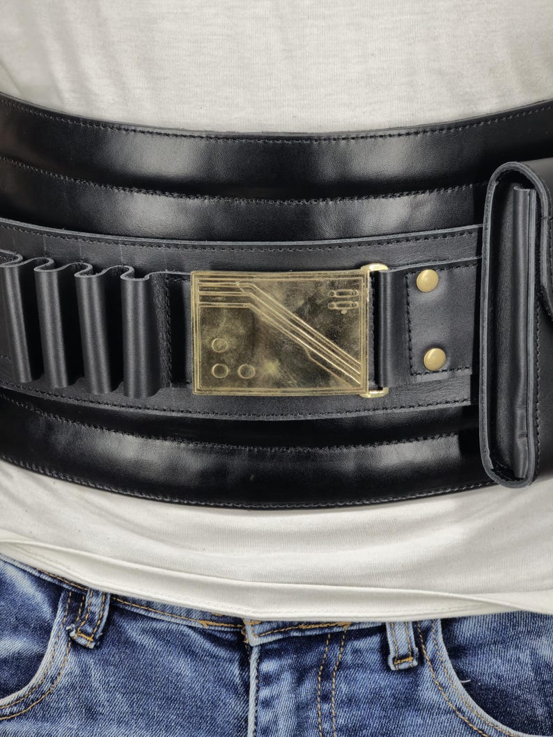 May include: Black leather belt with a gold-colored rectangular buckle. The belt features multiple loops and a small pouch. The buckle has a geometric design with three diagonal lines and three circles. The belt is worn over a white shirt and blue jeans.