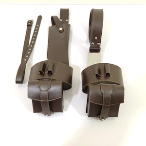 K&ouml;nnte beinhalten: Braunes Leder-Werkzeugg&uuml;rtel-Set mit zwei Taschen, einem G&uuml;rtel und einer Scheide. Die Taschen haben einen Klappenverschluss und sind an einem breiten G&uuml;rtel befestigt. Das Set ist zum Tragen von Gegenst&auml;nden konzipiert.