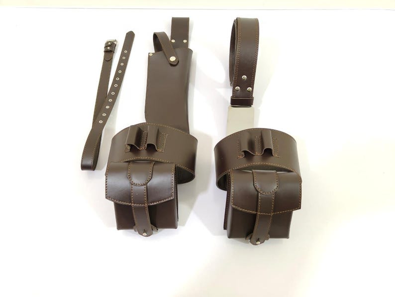 K&ouml;nnte beinhalten: Braunes Leder-Werkzeugg&uuml;rtel-Set mit zwei Taschen, einem G&uuml;rtel und einer Scheide. Die Taschen haben einen Klappenverschluss und sind an einem breiten G&uuml;rtel befestigt. Das Set ist zum Tragen von Gegenst&auml;nden konzipiert.