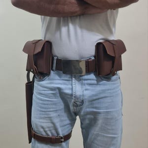 K&ouml;nnte beinhalten: Brauner Leder-Utility-G&uuml;rtel mit zwei Taschen, einem Holster und Beingurten. Der G&uuml;rtel hat eine silberne Schnalle und wird mit einer blauen Jeans und einem wei&szlig;en T-Shirt mit dem Aufdruck "IMPOSSIBLE" getragen.