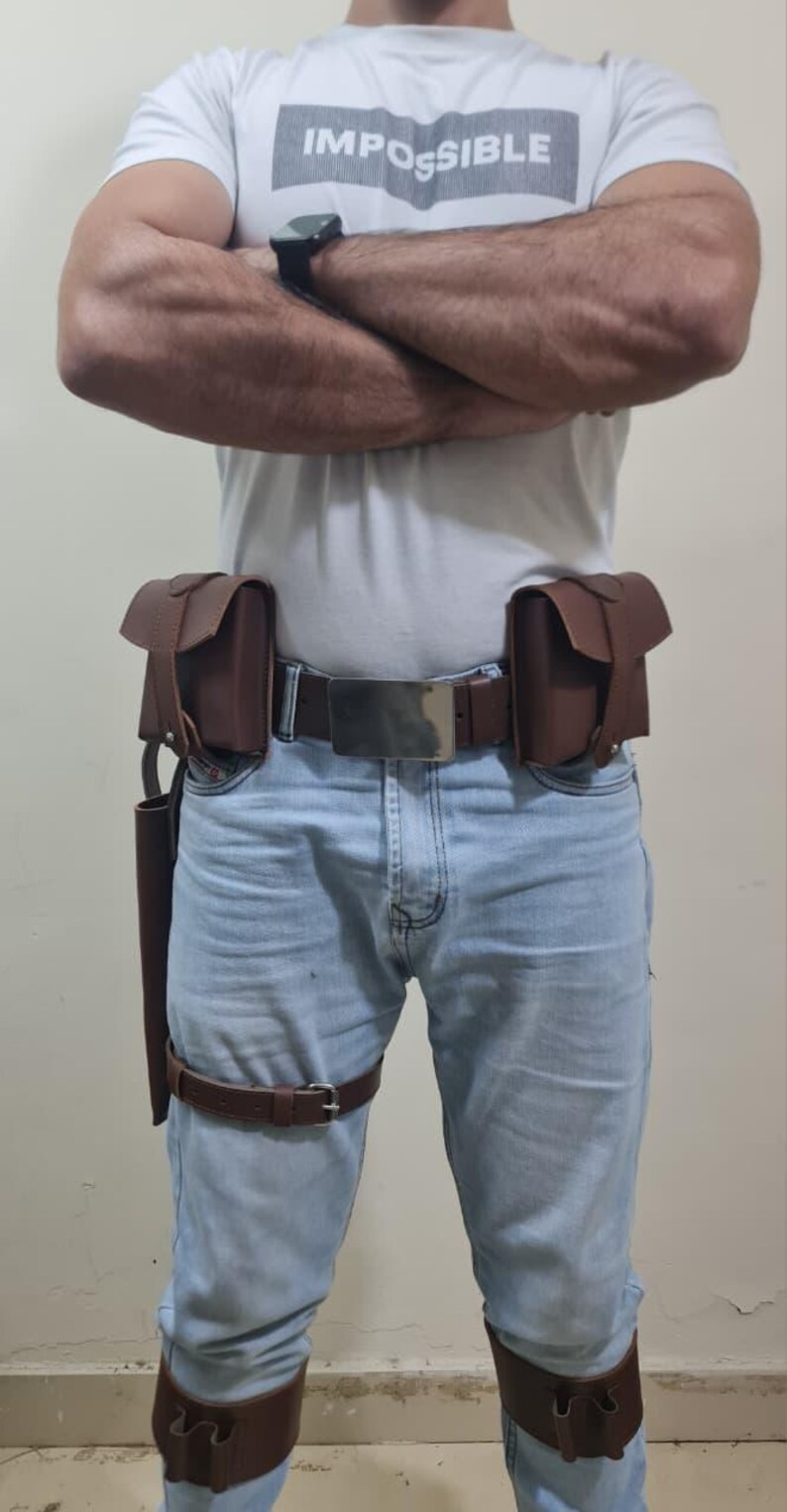 K&ouml;nnte beinhalten: Brauner Leder-Utility-G&uuml;rtel mit zwei Taschen, einem Holster und Beingurten. Der G&uuml;rtel hat eine silberne Schnalle und wird mit einer blauen Jeans und einem wei&szlig;en T-Shirt mit dem Aufdruck "IMPOSSIBLE" getragen.