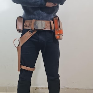 May include: A tan leather utility belt with multiple pouches and a holster, worn with black trousers. The belt features a silver buckle with the letter "D" and other geometric designs. An orange accent is visible on one of the pouches.