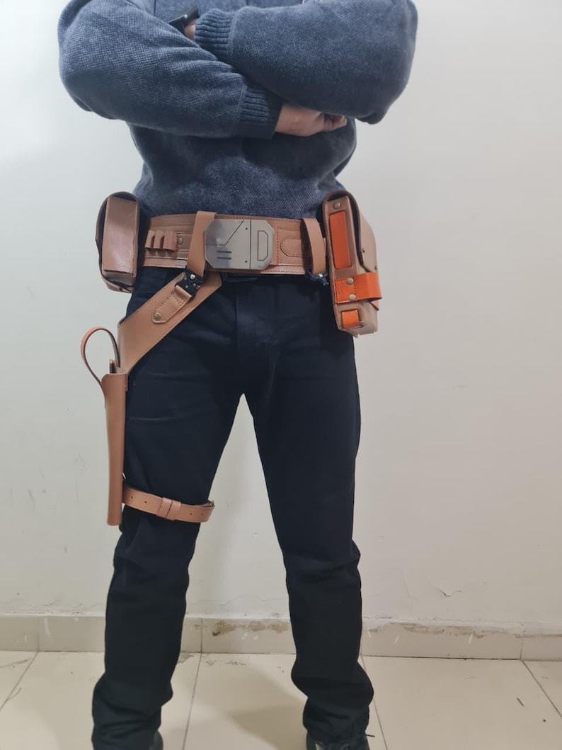 May include: A tan leather utility belt with multiple pouches and a holster, worn with black trousers. The belt features a silver buckle with the letter "D" and other geometric designs. An orange accent is visible on one of the pouches.