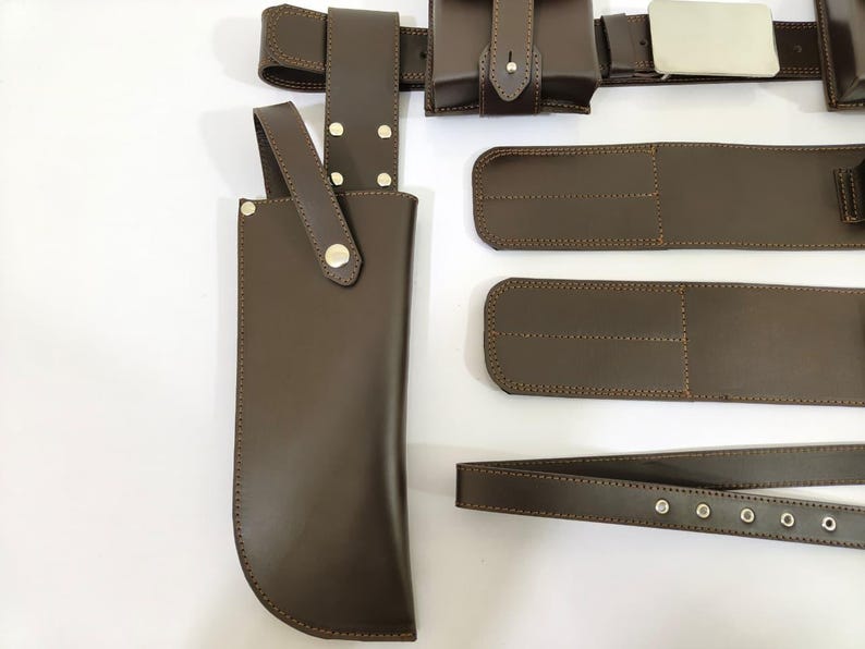 K&ouml;nnte beinhalten: Ein braunes Lederg&uuml;rtel- und Zubeh&ouml;rset. Das Set enth&auml;lt einen G&uuml;rtel mit silberner Schnalle, Taschen und eine Messerscheide. Die Artikel sind mit einem Kontrastfaden gen&auml;ht. Der Hintergrund ist wei&szlig;.