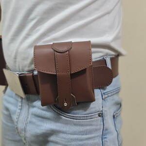 K&ouml;nnte beinhalten: Eine braune Ledertasche, die an einem braunen G&uuml;rtel befestigt ist und &uuml;ber einer blauen Jeans getragen wird. Die Tasche hat einen Klappenverschluss mit einem gen&auml;hten Design und einem silberfarbenen Niet. Der G&uuml;rtel hat eine silberfarbene Schnalle.