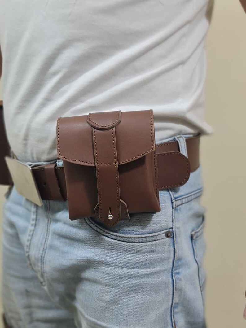 K&ouml;nnte beinhalten: Eine braune Ledertasche, die an einem braunen G&uuml;rtel befestigt ist und &uuml;ber einer blauen Jeans getragen wird. Die Tasche hat einen Klappenverschluss mit einem gen&auml;hten Design und einem silberfarbenen Niet. Der G&uuml;rtel hat eine silberfarbene Schnalle.