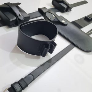 K&ouml;nnte beinhalten: Eine Sammlung schwarzer Lederaccessoires, darunter eine Handgelenksmanschette, ein G&uuml;rtel und Beutel. Die Artikel sind auf einer wei&szlig;en Oberfl&auml;che angeordnet, die die Details des Leders und der N&auml;hte zeigt. Das Set scheint f&uuml;r einen bestimmten Zweck bestimmt zu sein.