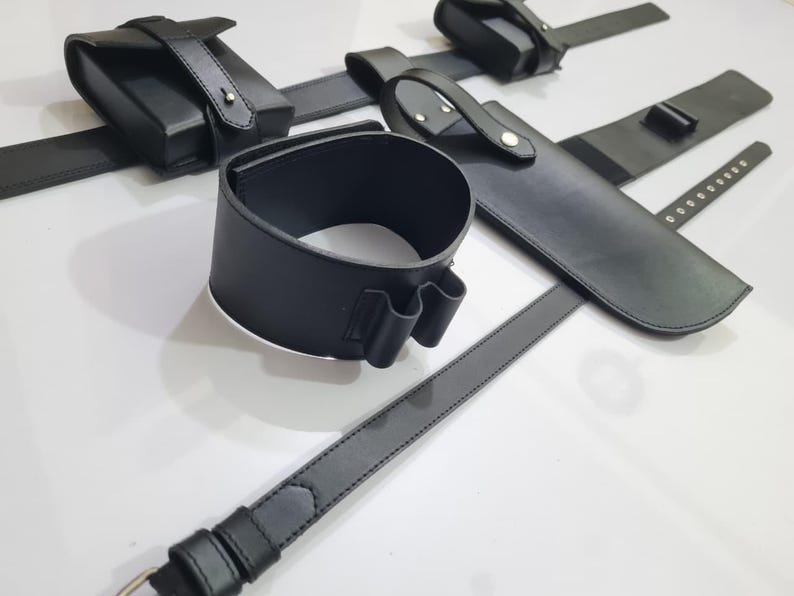 K&ouml;nnte beinhalten: Eine Sammlung schwarzer Lederaccessoires, darunter eine Handgelenksmanschette, ein G&uuml;rtel und Beutel. Die Artikel sind auf einer wei&szlig;en Oberfl&auml;che angeordnet, die die Details des Leders und der N&auml;hte zeigt. Das Set scheint f&uuml;r einen bestimmten Zweck bestimmt zu sein.