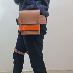 May include: A tan leather utility belt with a rectangular pouch and an orange strap. The belt is worn around the waist and has a matching leather strap around the thigh. The pouch has a flap closure and metal rivets.