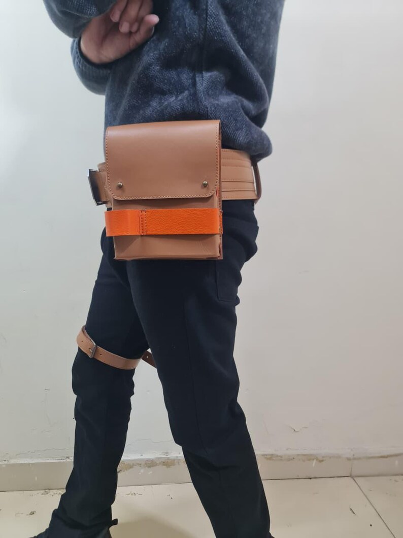 May include: A tan leather utility belt with a rectangular pouch and an orange strap. The belt is worn around the waist and has a matching leather strap around the thigh. The pouch has a flap closure and metal rivets.