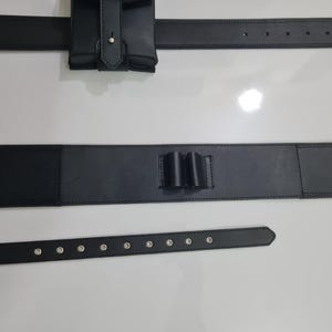 K&ouml;nnte beinhalten: Schwarzer Leder-Utility-G&uuml;rtel mit mehreren Komponenten. Der G&uuml;rtel umfasst eine Tasche, einen Abschnitt mit drei Schlaufen und zwei separate G&uuml;rtel. Die G&uuml;rtel haben silberfarbene Nieten. Die Artikel werden auf einer wei&szlig;en Oberfl&auml;che pr&auml;sentiert.