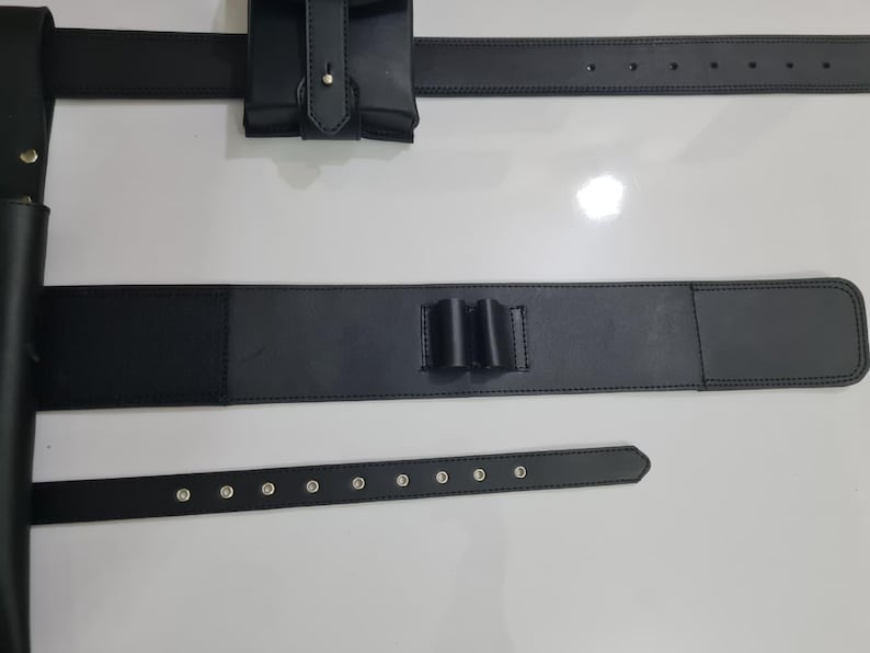 K&ouml;nnte beinhalten: Schwarzer Leder-Utility-G&uuml;rtel mit mehreren Komponenten. Der G&uuml;rtel umfasst eine Tasche, einen Abschnitt mit drei Schlaufen und zwei separate G&uuml;rtel. Die G&uuml;rtel haben silberfarbene Nieten. Die Artikel werden auf einer wei&szlig;en Oberfl&auml;che pr&auml;sentiert.