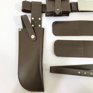 K&ouml;nnte beinhalten: Ein braunes Lederg&uuml;rtel- und Zubeh&ouml;rset. Das Set enth&auml;lt einen G&uuml;rtel mit silberner Schnalle, Taschen und eine Messerscheide. Die Artikel sind aus braunem Leder mit dekorativen N&auml;hten und silbernen Nieten gefertigt.