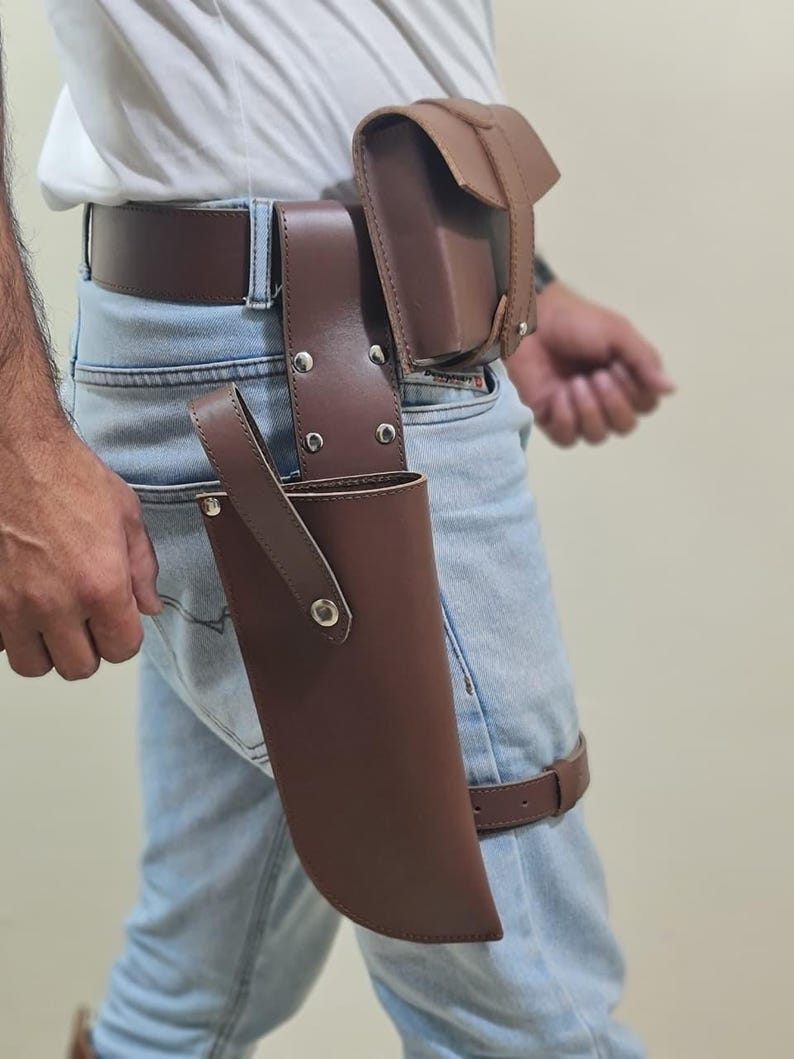 K&ouml;nnte beinhalten: Brauner Leder-Utility-G&uuml;rtel mit Holster, Tasche und Beingurt. Der G&uuml;rtel hat silberne Nieten und eine Schlaufe f&uuml;r ein Messer oder Werkzeug. Das Design eignet sich f&uuml;r Cosplay oder einen robusten, funktionalen Stil.