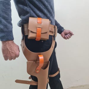 May include: A tan leather holster and belt set with orange accents. The set includes a belt with a pouch, a thigh holster, and straps. The leather is a light brown colour, and the accents are bright orange.