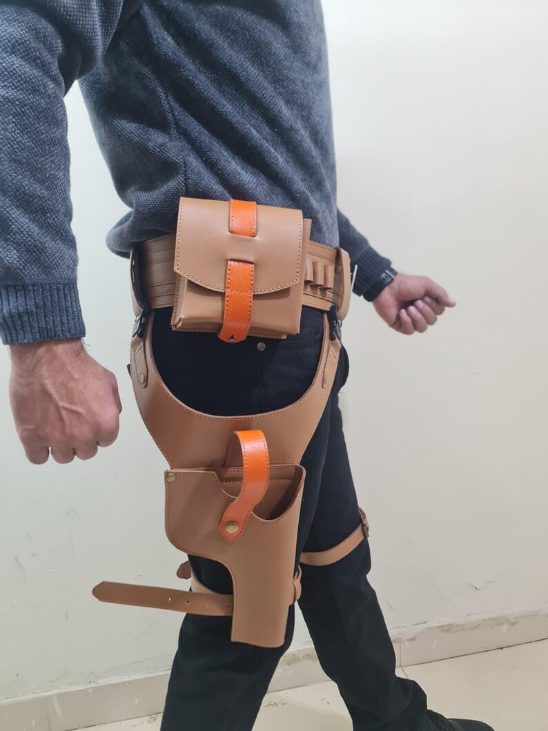 May include: A tan leather holster and belt set with orange accents. The set includes a belt with a pouch, a thigh holster, and straps. The leather is a light brown colour, and the accents are bright orange.