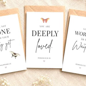 May include: Three white greeting cards with inspirational quotes and butterfly illustrations. The cards are in brown envelopes and surrounded by small white flowers. The text includes "God's Not Done", "You Are Deeply Loved", and "God Is Working".