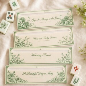 Op de afbeelding: Vier rechthoekige, gebroken witte Mahjong-tegelhouders met groene bloemillustraties en tekst. De houders tonen de zinnen Joy Is Always in the Game, Here's to Lucky Draws, Winning Hands en A Beautiful Day to Mahj, samen met losse speelstenen.