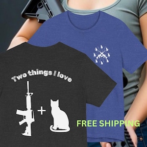 May include: A dark grey t-shirt featuring the white text Two things I love, a white rifle silhouette, a plus sign, and a white cat silhouette. A blue t-shirt with a white graphic of a rifle surrounded by small cat silhouettes is also visible.