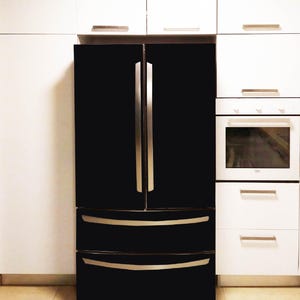 K&ouml;nnte beinhalten: Ein schwarzer French-Door-K&uuml;hlschrank mit zwei vertikalen T&uuml;ren und zwei unteren Gefrierschubladen, ausgestattet mit langen, schmalen, silberfarbenen Metallgriffen. Das Ger&auml;t ist zwischen wei&szlig;en K&uuml;chenschr&auml;nken und einem Backofen platziert.