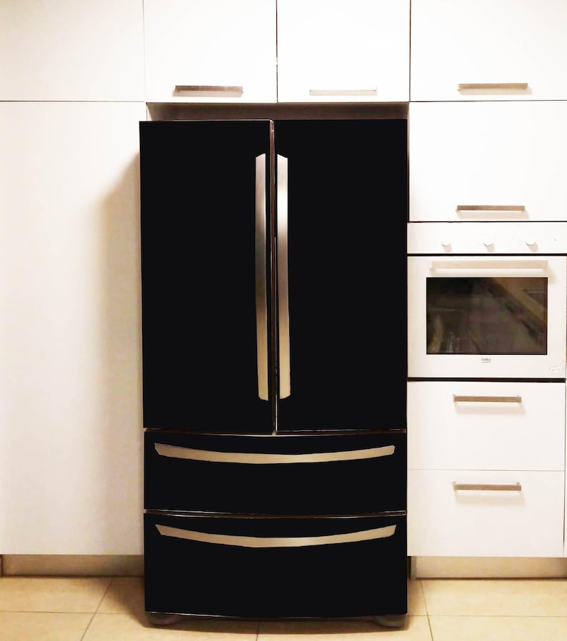 K&ouml;nnte beinhalten: Ein schwarzer French-Door-K&uuml;hlschrank mit zwei vertikalen T&uuml;ren und zwei unteren Gefrierschubladen, ausgestattet mit langen, schmalen, silberfarbenen Metallgriffen. Das Ger&auml;t ist zwischen wei&szlig;en K&uuml;chenschr&auml;nken und einem Backofen platziert.