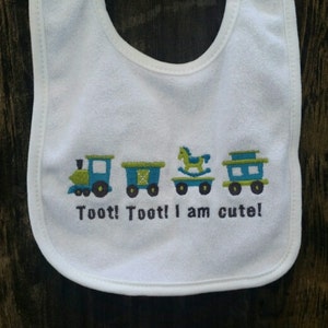 May include: White baby bib with a train, rocking horse, and train car design. The bib has the text "Toot! Toot! I am cute!" embroidered on it.