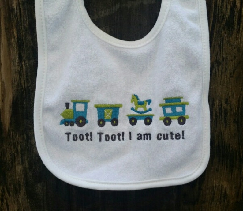 May include: White baby bib with a train, rocking horse, and train car design. The bib has the text "Toot! Toot! I am cute!" embroidered on it.