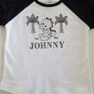 Puede incluir: Camiseta blanca y negra de estilo ragl&aacute;n con un dise&ntilde;o de dinosaurio y palmeras. El nombre "JOHNNY" est&aacute; bordado debajo del gr&aacute;fico. La camiseta est&aacute; etiquetada como "Creations of Grace 2T".