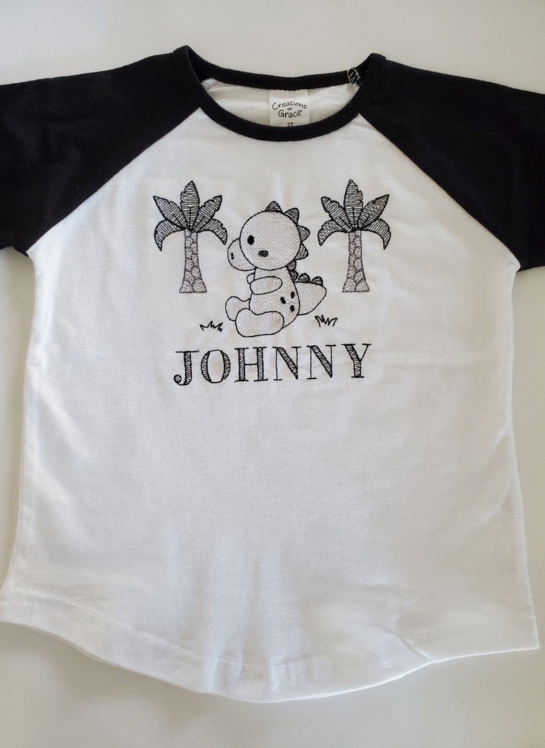 Puede incluir: Camiseta blanca y negra de estilo ragl&aacute;n con un dise&ntilde;o de dinosaurio y palmeras. El nombre "JOHNNY" est&aacute; bordado debajo del gr&aacute;fico. La camiseta est&aacute; etiquetada como "Creations of Grace 2T".