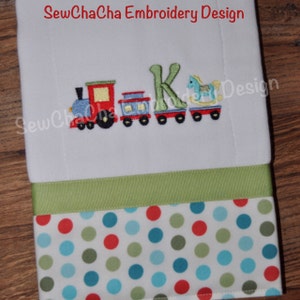 May include: Embroidered design featuring a red, blue, and green toy train, a rocking horse, and the letter K. The design is on a white fabric with a green ribbon and a red, green, and blue polka dot fabric border.