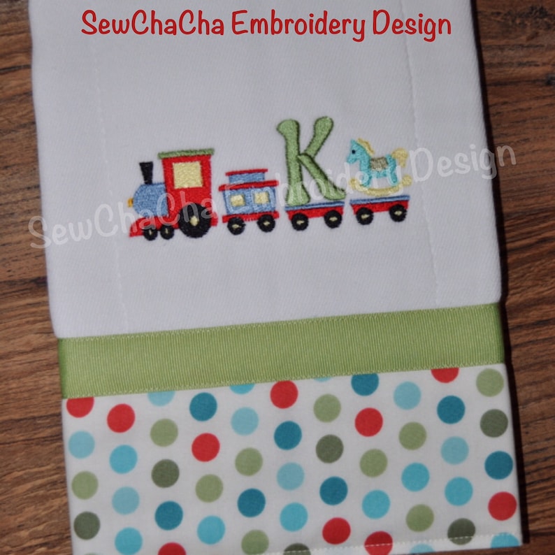 May include: Embroidered design featuring a red, blue, and green toy train, a rocking horse, and the letter K. The design is on a white fabric with a green ribbon and a red, green, and blue polka dot fabric border.