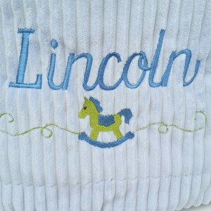 May include: A light blue corduroy pillow with the name "Lincoln" embroidered in blue thread. A green rocking horse is embroidered below the name.