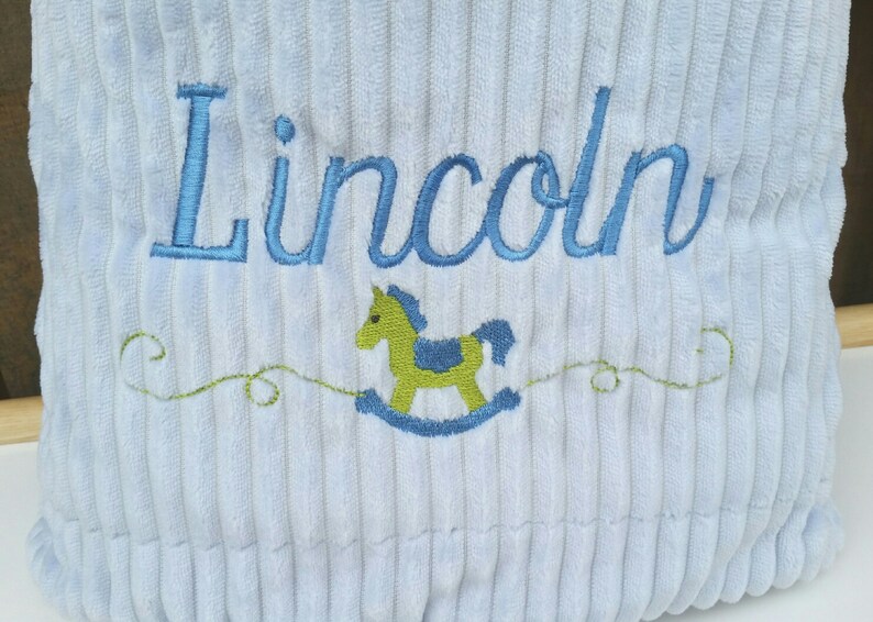 May include: A light blue corduroy pillow with the name "Lincoln" embroidered in blue thread. A green rocking horse is embroidered below the name.