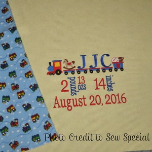 May include: A yellow baby blanket with a blue and white train pattern border. The blanket is embroidered with a train carrying the letters JJC, the numbers 2 pounds 13 ounces and 14 inches, and the date August 20, 2016.