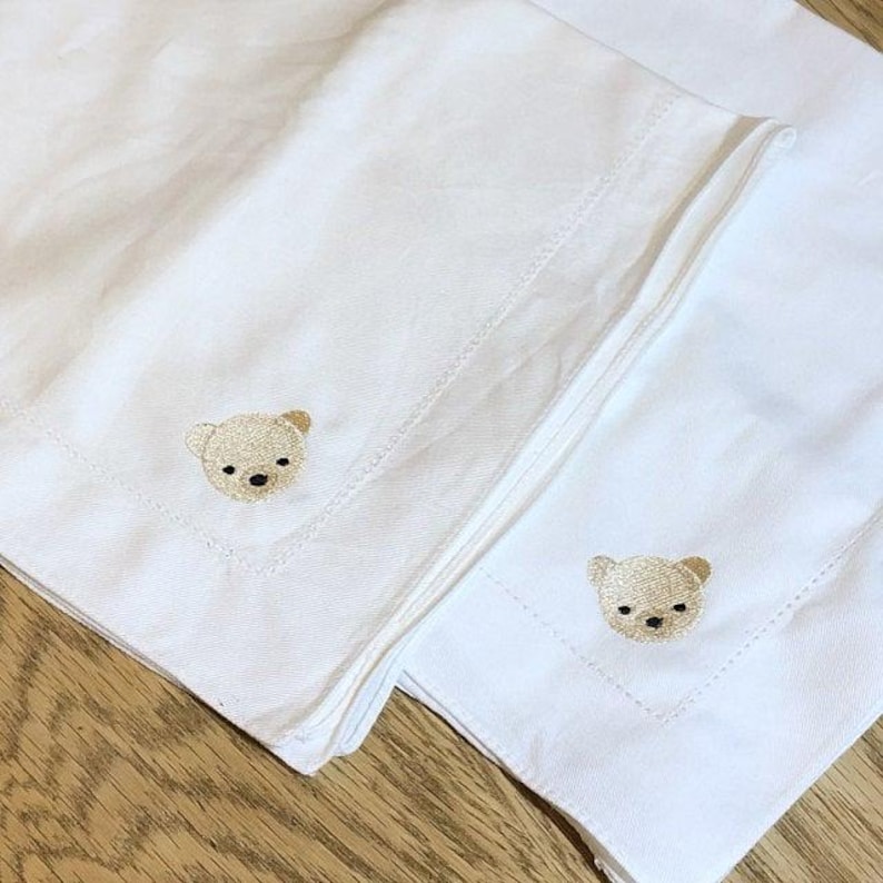 以下が含まれることがあります： 刺繍された茶色のクマの顔がついた白いリネンナプキンが2枚。ナプキンは半分に折り畳まれ、重ねられています。