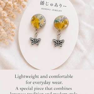 以下が含まれることがあります： 丸い黄色と青色の樹脂トップとシルバーの蝶のチャームが付いたイヤリング。イヤリングは、日本語のテキストと「軽量で快適な普段使い」という言葉が書かれた白いカードに表示されています。