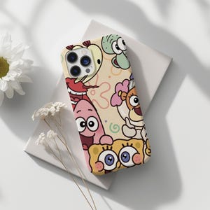 Puede incluir: Funda de teléfono con personajes de dibujos animados sobre un fondo beige claro. La funda tiene un diseño colorido con personajes como Bob Esponja y Patricio Estrella. La funda está sobre una superficie blanca.