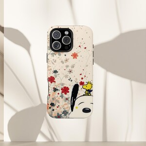 May include: A phone case with a floral pattern in red and white, and a cartoon illustration of Snoopy and Woodstock. The case has a cream background and a black camera module.