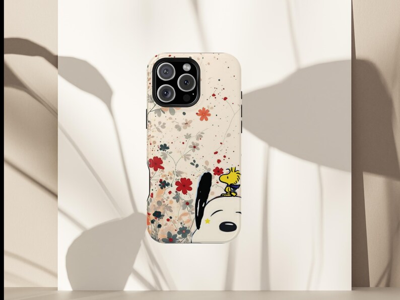 May include: A phone case with a floral pattern in red and white, and a cartoon illustration of Snoopy and Woodstock. The case has a cream background and a black camera module.