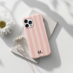 Puede incluir: Una funda de teléfono a rayas rosas y crema con las iniciales "SL" en negro. La funda está sobre una superficie blanca, con una flor blanca y ramitas de flores secas cerca.